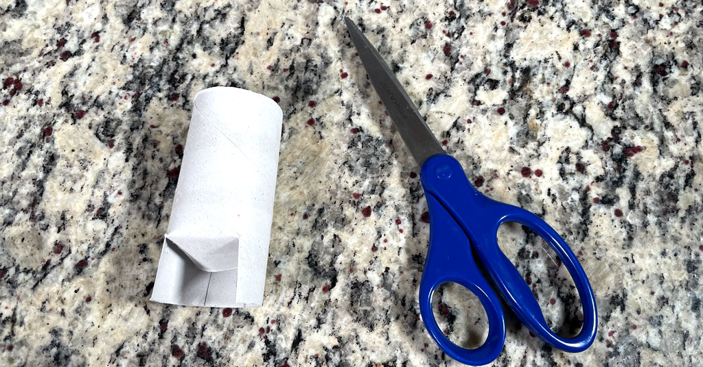 One toilet paper roll with no toilet paper with four slits cut into the side to make a pot with a pair of scissors with blue handles.