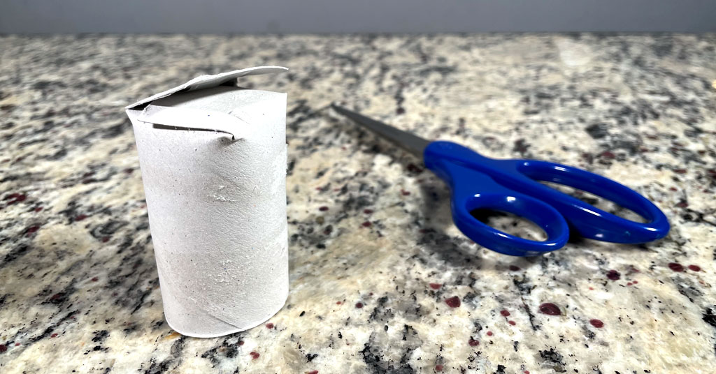 One toilet paper roll without toilet paper on it with the bottom folded into a pot and a pair of scissors with blue handles