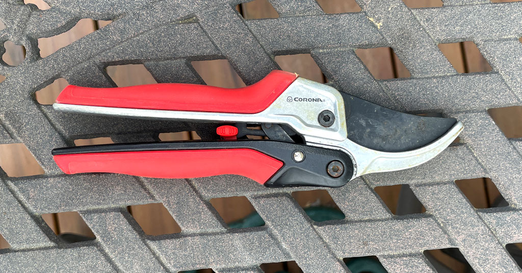 Red handled garden pruners sitting on a metal table. Garden pruners made by Corona Tools.