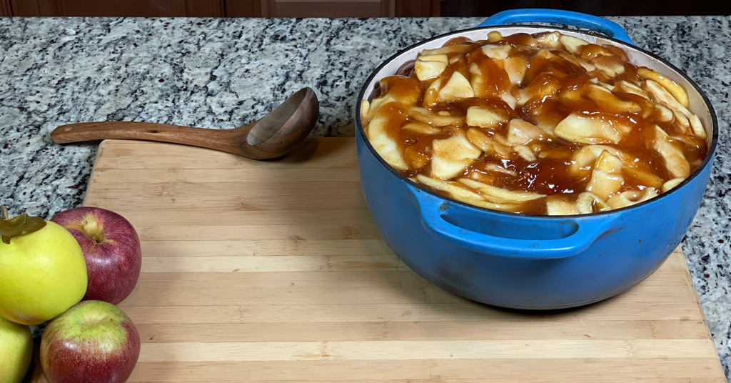 Apple Pie Filling in a blue enameled dutch oven sitting on a cutting board with a wooden ladle.
