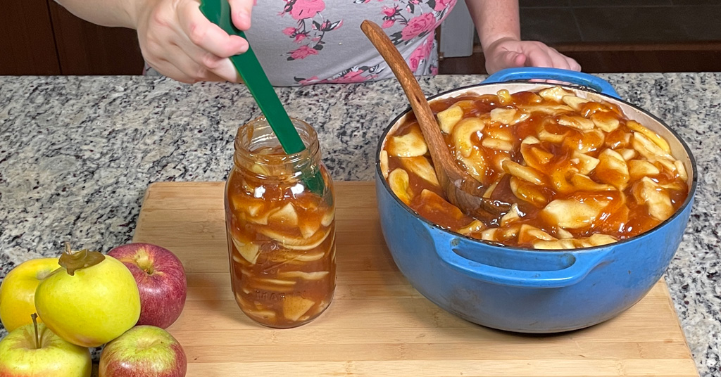 Apple Pie Filling in a blue enameled dutch oven. Some filling has been ladled into a quart size canning jar. Green de-bubbler being used to remove bubbles from apple pie filling