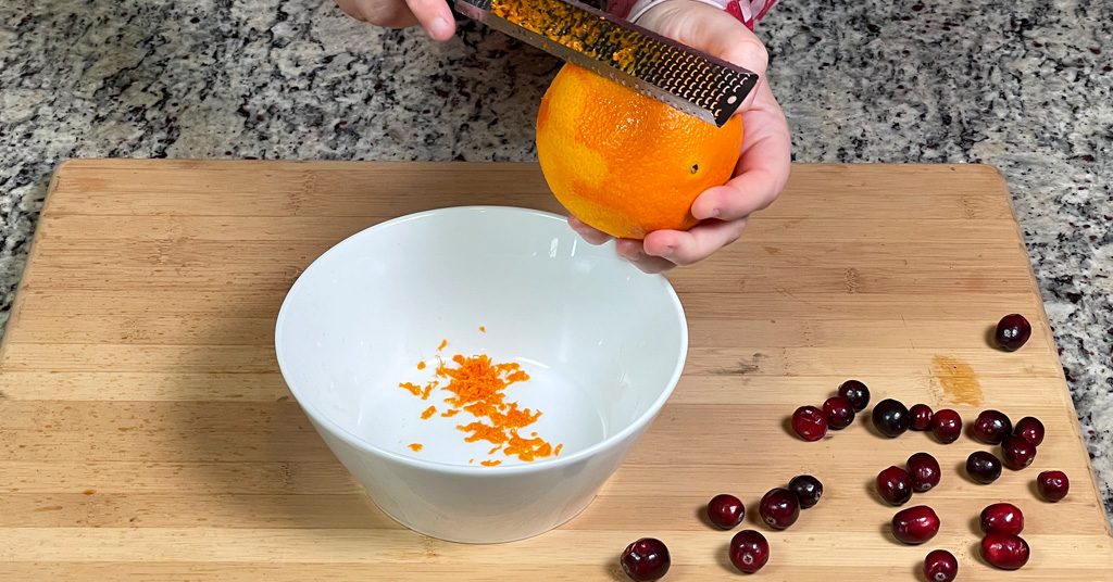White bowl sitting on a wooden cutting board. Hands zesting an orange into the white bowl.
