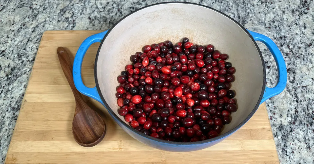 Fresh Cranberries in a blue enameled dutch oven sitting on a wooden cutting board with a wooden spoon.