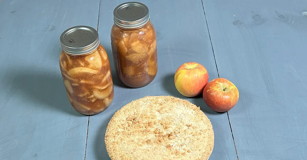 Two quart sized mason jars of Apple Pie Filling that have been water bath canned. Apple pie made with apple pie filling with two red and yellow apples sitting on a blue background.