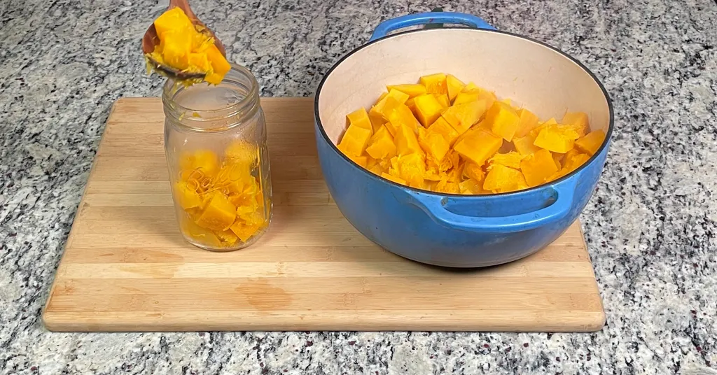 Blue enameled dutch oven with hot pumpkin cubes being ladled into a quart sized mason jar.