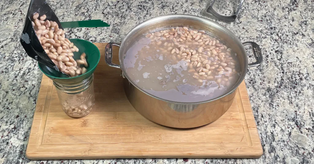 Canning Pinto Beans with a Pressure Canner. Ladling hot pinto beans into a hot pint sized mason jar next to a stainless steal pot full of pinto beans.