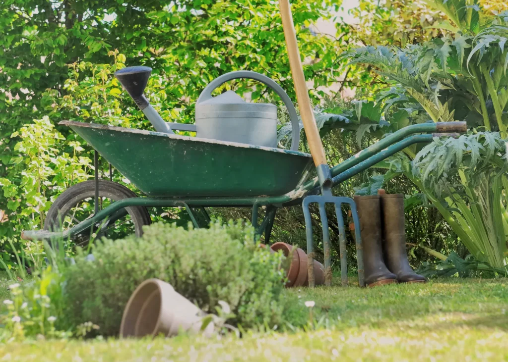 Green wheelbarrow, watering can, garden fork, post and boots sitting in a garden.